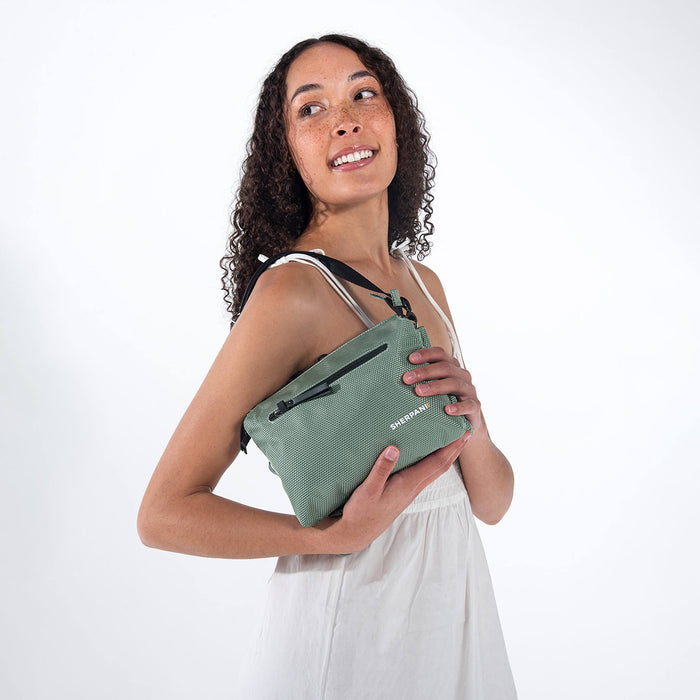 Woman holding a Sherpani Mei Aloe Mist Green Anti-theft Travel Trio Crossbody by short strap against white backdrop.   #color_aloe-mist-green