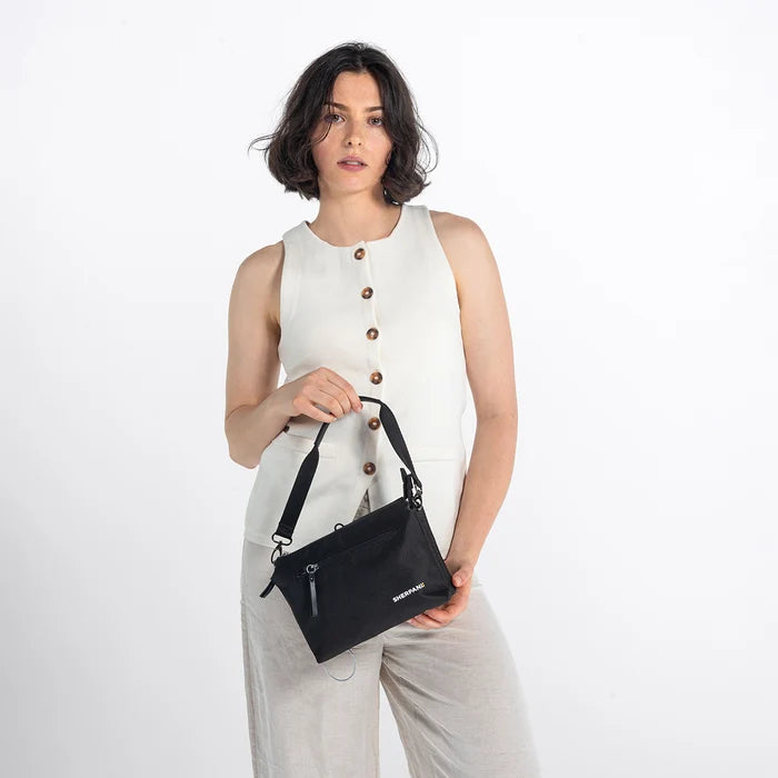 Woman holding a Sherpani Mei Carbon Black Anti-theft Travel Trio Crossbody by the short strap against white backdrop. #color_carbon-black