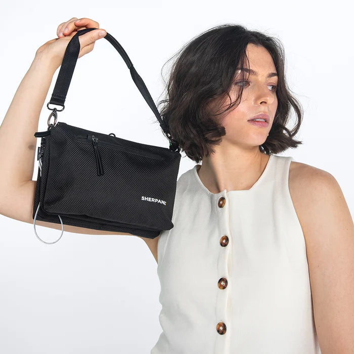 Woman holding a Sherpani Mei Carbon Black Anti-theft Travel Trio Crossbody by the short strap against white backdrop. #color_carbon-black