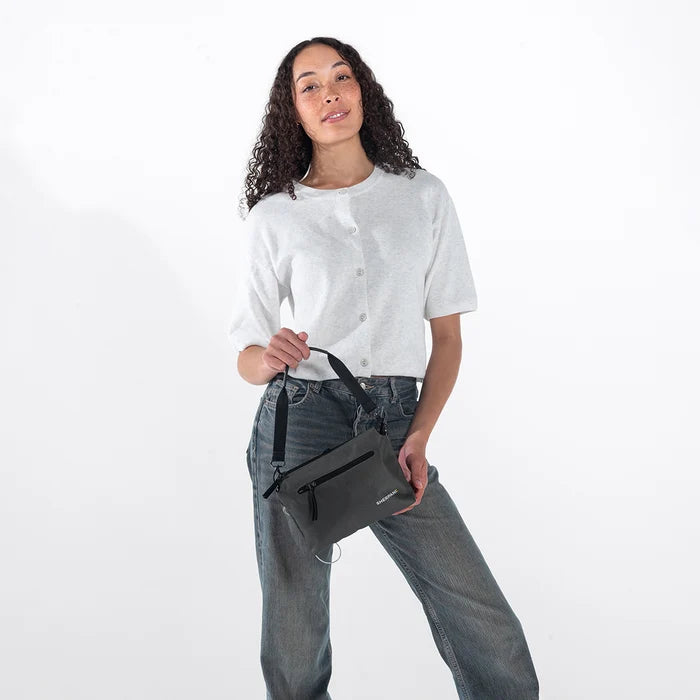 Woman holding a Sherpani Mei Granite Anti-theft Travel Trio Crossbody by the short strap against white backdrop. #color_granite-grey