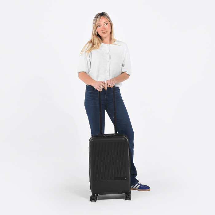 Woman holding a Sherpani Meridian Black Carry-On Luggage by the handle against white backdrop. #color_black