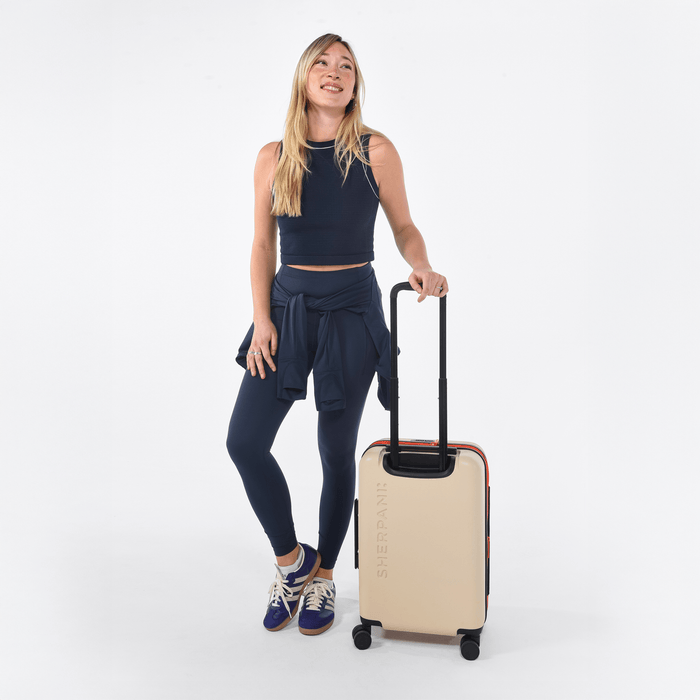 Woman holding a Sherpani Meridian Chai Beige Carry-On Luggage by the handle against white backdrop. #color_chai-beige