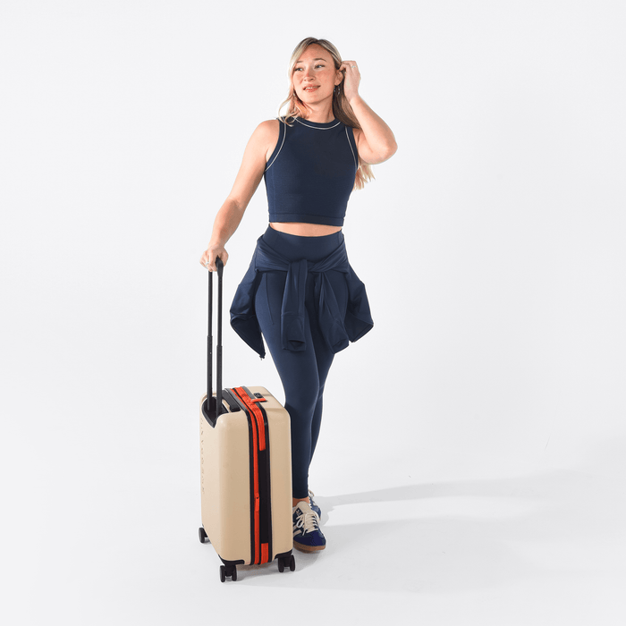 Woman holding a Sherpani Meridian Chai Beige Carry-On Luggage by the handle against white backdrop. #color_chai-beige