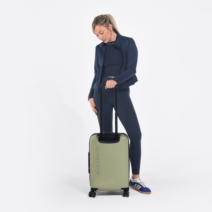 Woman holding a Sherpani Meridian Matcha Carry-On Luggage by the handle against white backdrop. #color_matcha-green