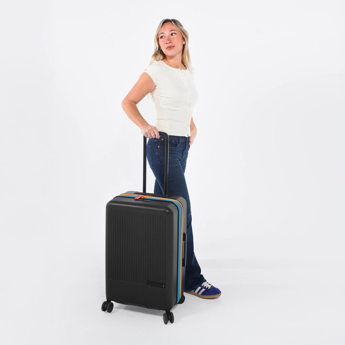 Woman holding a Sherpani Meridian Chromatic Black Checked-Luggage by the handle against white backdrop #color_chromatic-black