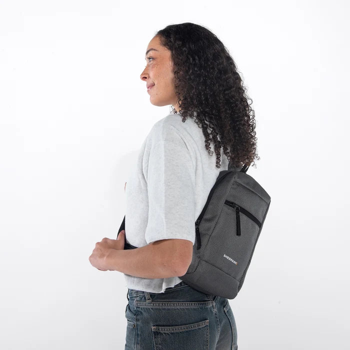 Woman wearing a Sherpani Metro Granite Anti-theft Reversible Sling on her back against white backdrop. #color_granite-grey