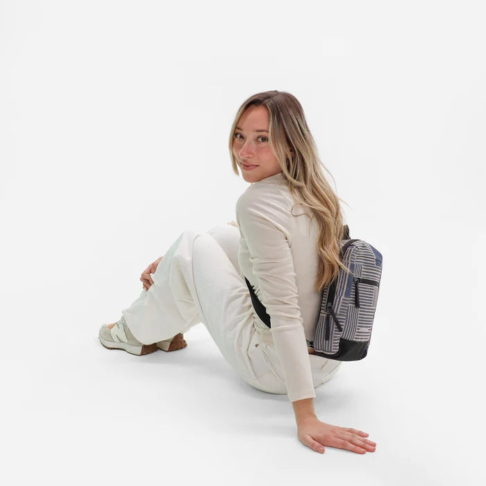 Woman sitting with a Sherpani Metro Passport Anti-theft Reversible Sling on her back against white backdrop. #color_passport-print