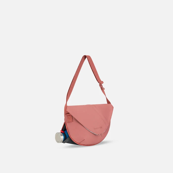 A Sherpani Milli Desert Pink Mini Messenger, 3/4 view showing front flap pocket and water bottle pocket on white backdrop.   #color_desert-pink