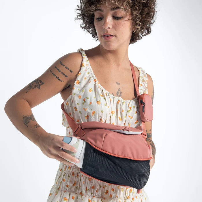 Woman pulling a water bottle out of a Sherpani Milli Desert Pink Mini Messenger against white backdrop.  #color_desert-pink