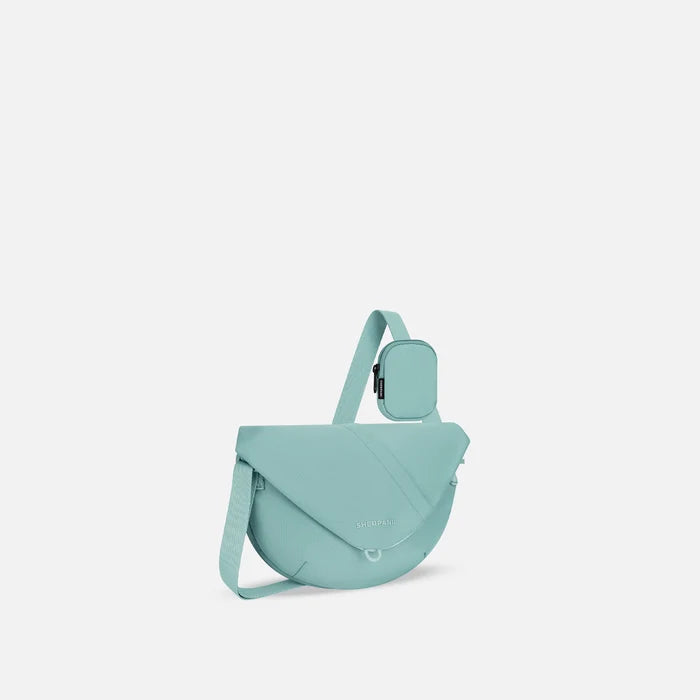 A Sherpani Milli Surf Blue Mini Messenger, 3/4 view showing front flap pocket and strap on white backdrop.  #color_surf-blue