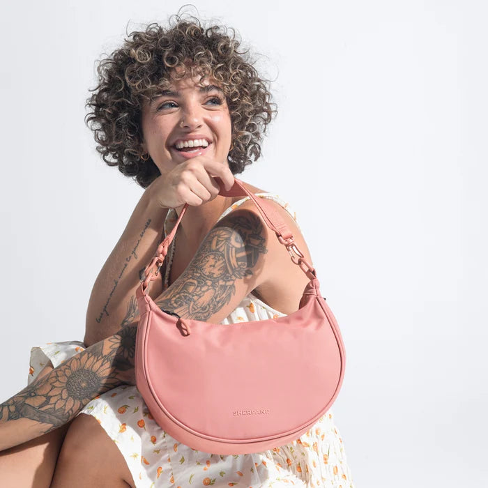 A woman holding a Sherpani Mina Desert Pink Medium Crossbody Bag in her hands with short-strap attachment. #color_desert-pink