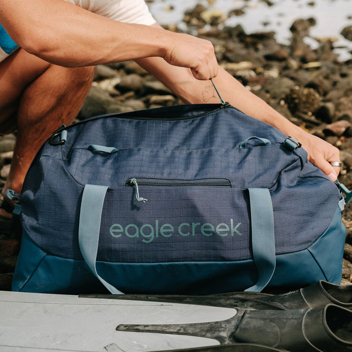 A person unzipping a navy blue duffle bag on a rocky beach