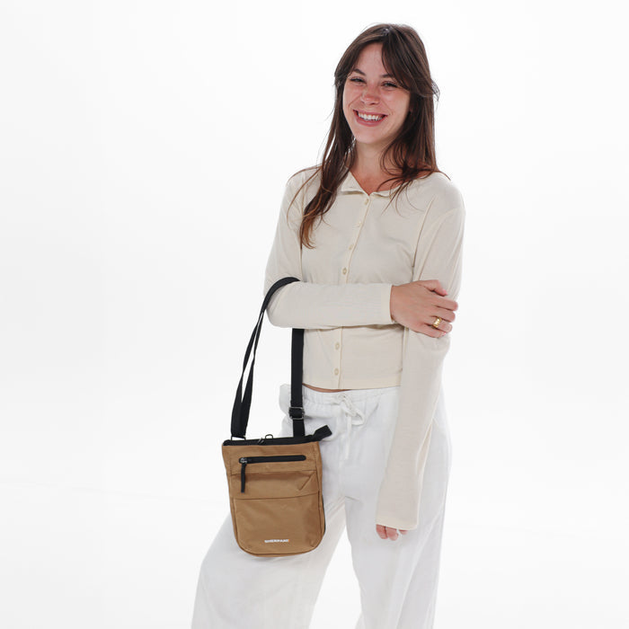 Woman holding a Sherpani Prima Sand Anti-Theft Travel Crossbody by long strap against white background.  #color_sand-tan
