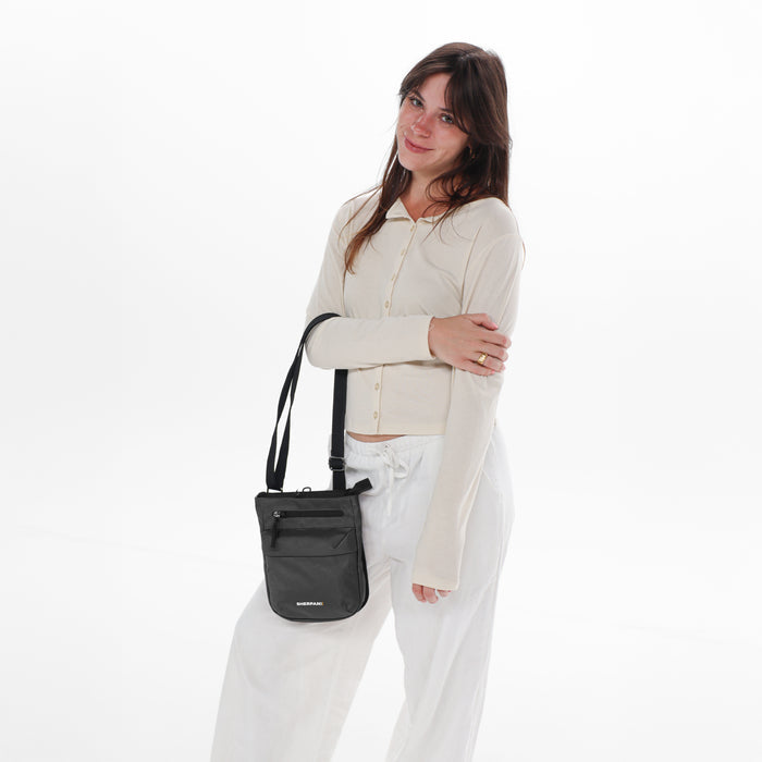 Woman holding a Sherpani Prima Granite Anti-Theft Travel Crossbody by long strap against white background.  #color_granite-grey