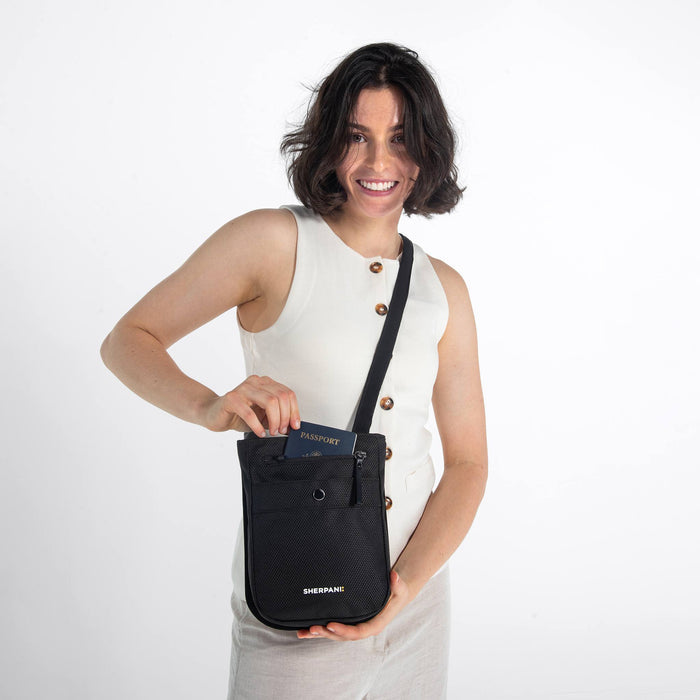 Woman wearing a Sherpani Prima Carbon Black Travel Crossbody pulls out a passport out of bag against white background. #color_carbon-black