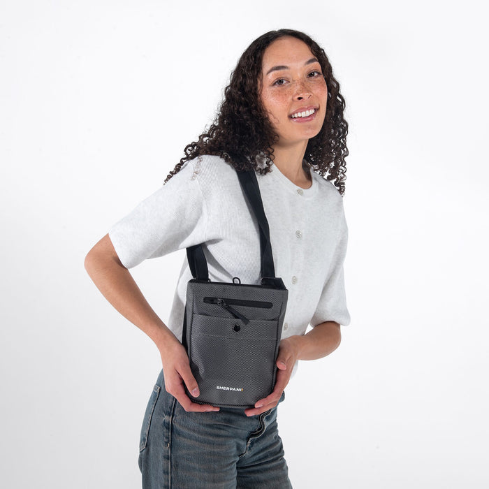 Woman wearing a Sherpani Prima Granite Travel Crossbody on her shoulder against white background.  #color_granite-grey
