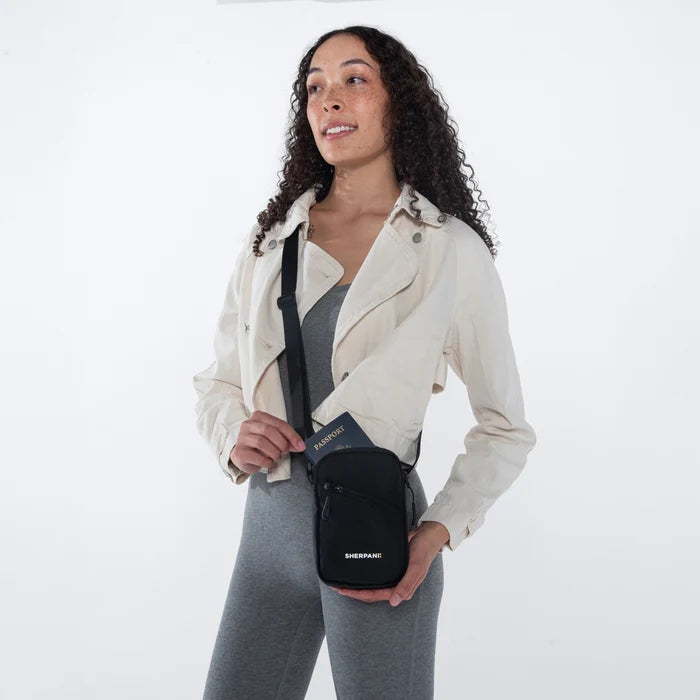 Woman placing a passport into a Sherpani Rogue Raven Black Mini Crossbody on her shoulder against white background.   #color_raven-black
