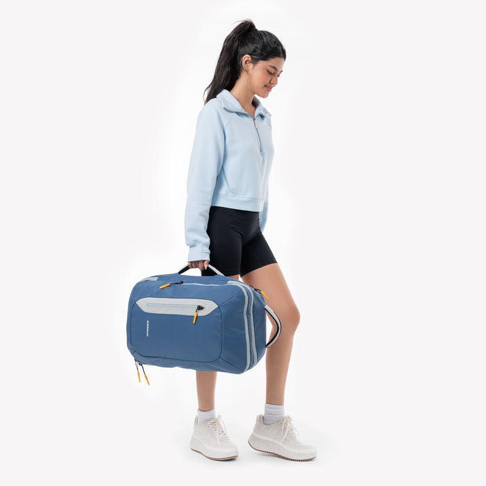 Woman holding a Sherpani Santiago Seastone Travel Backpack by side handle against white backdrop. #color_seastone