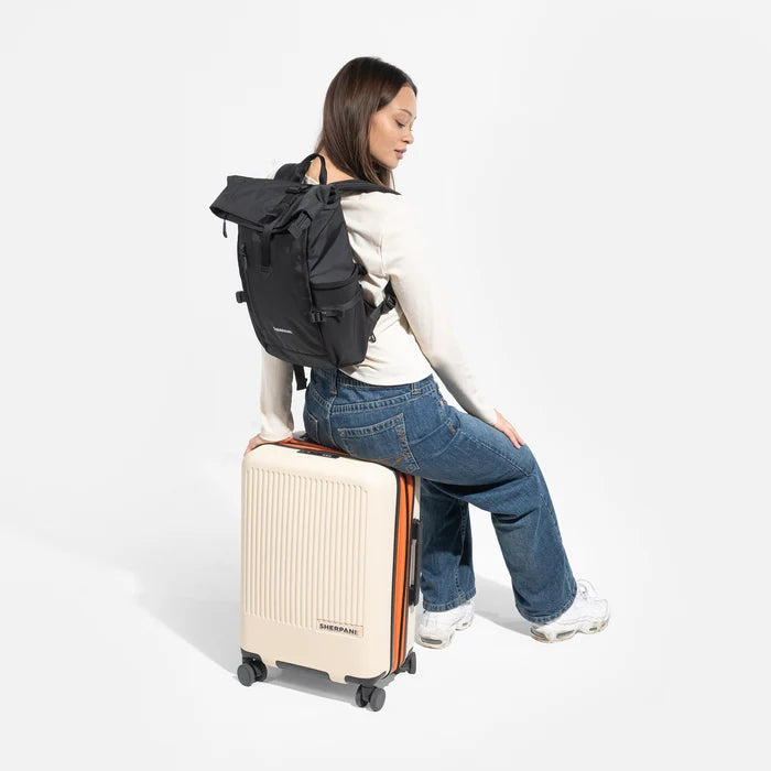 A woman sitting on a Sherpani Meridian Luggage with a Sherpani Sedona Raven Black Backpack on her back against white background. #color_raven-black