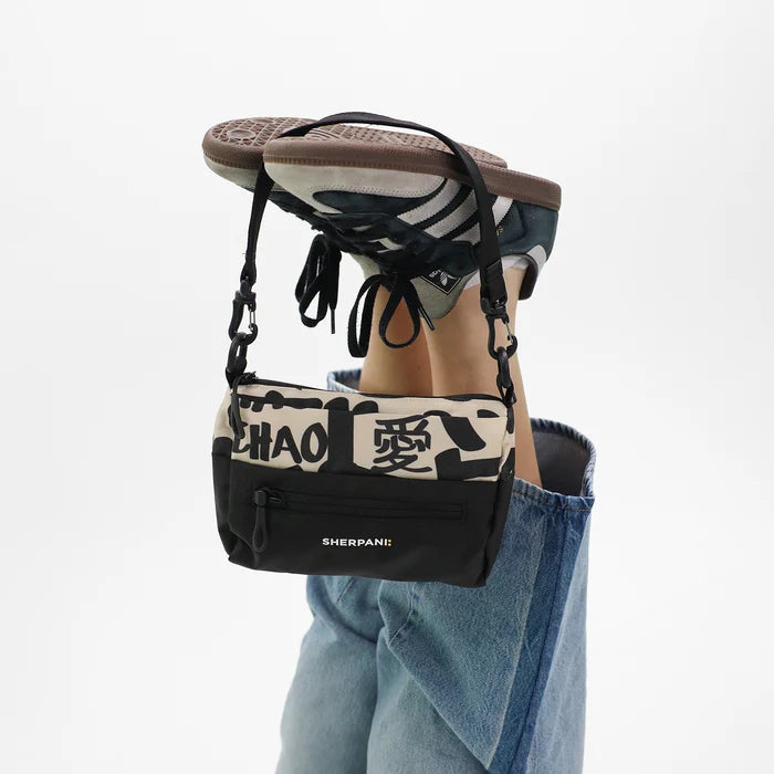 A Sherpani Skye Ciao Shoulder Bag hanging by a woman's shoes against white backdrop. #color_ciao-print