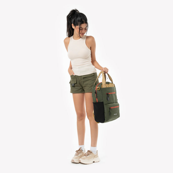 Woman holding a Sherpani Sojourn Antelope Multi-Wear Pack by handheld straps against white backdrop. #color_antelope