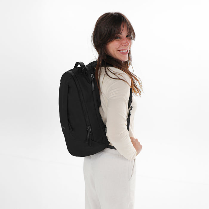 A woman wearing a Sherpani Solstice Carbon Black Anti-Theft Travel Backpack on her back against a white background.   #color_carbon-black