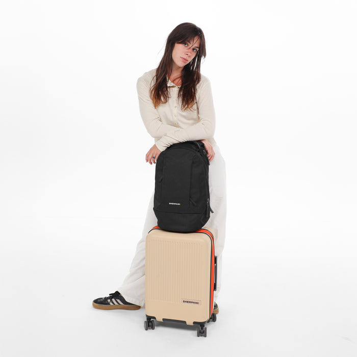 A woman holding a Sherpani Solstice Carbon Black Anti-Theft Travel Backpack on top of a Sherpani Meridian Luggage. #color_carbon-black