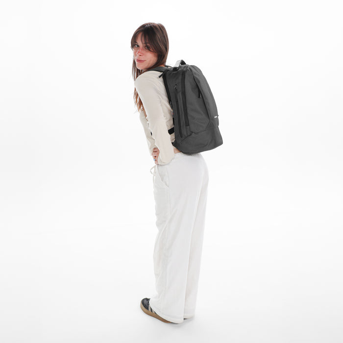 A woman wearing a Sherpani Solstice Granite Anti-Theft Travel Backpack on her back against a white background.  #color_granite-grey