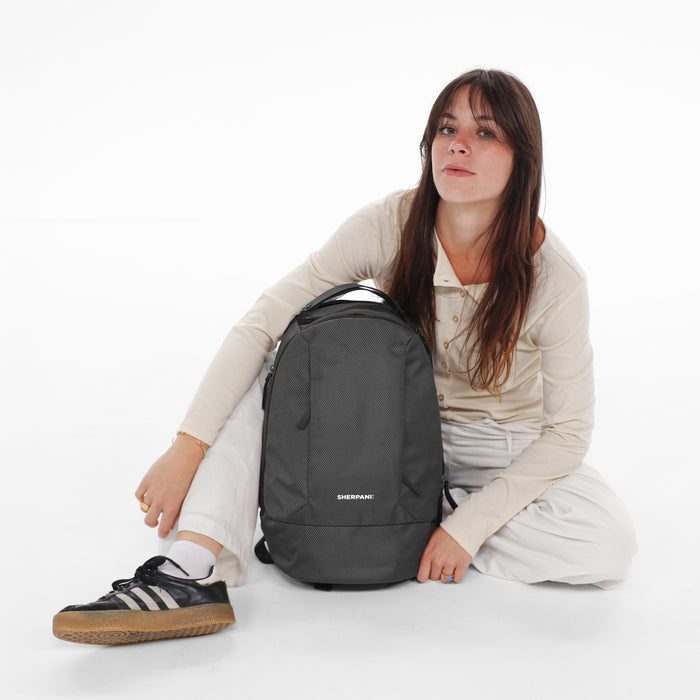 A woman sitting next to a Sherpani Solstice Granite Anti-Theft Travel Backpack against a white background.   #color_granite-grey