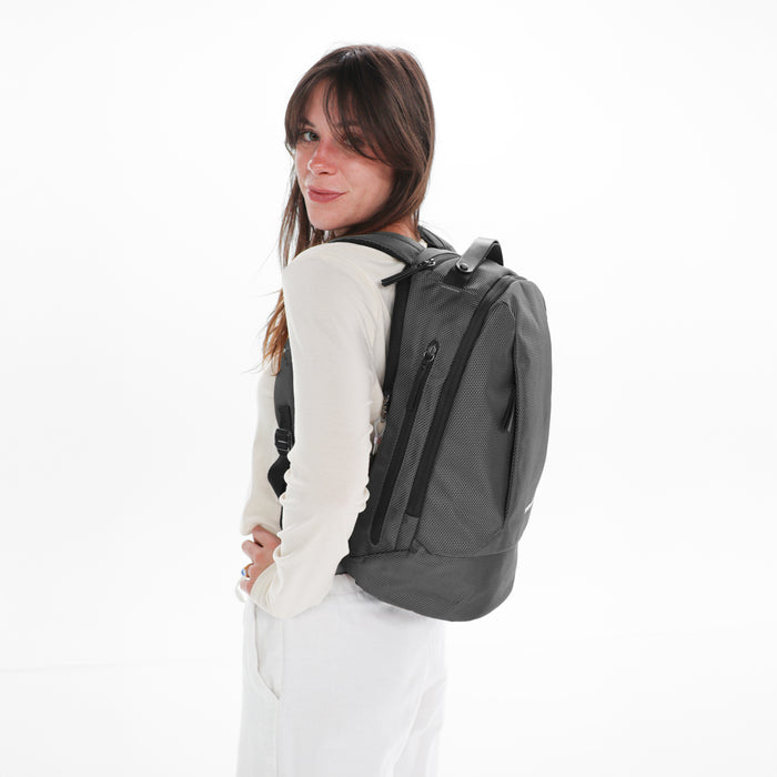 A woman wearing a Sherpani Solstice Granite Anti-Theft Travel Backpack on her back against a white background.  #color_granite-grey