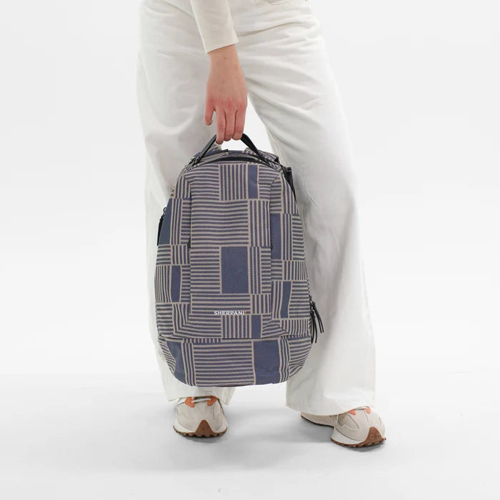 A woman holding a Sherpani Solstice Smoky Orchid Anti-Theft Travel Backpack by handheld strap against a white background.  #color_passport-print