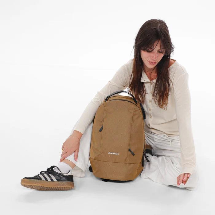 A woman sitting next to a Sherpani Solstice Sand Anti-Theft Travel Backpack against a white background.  #color_sand-tan
