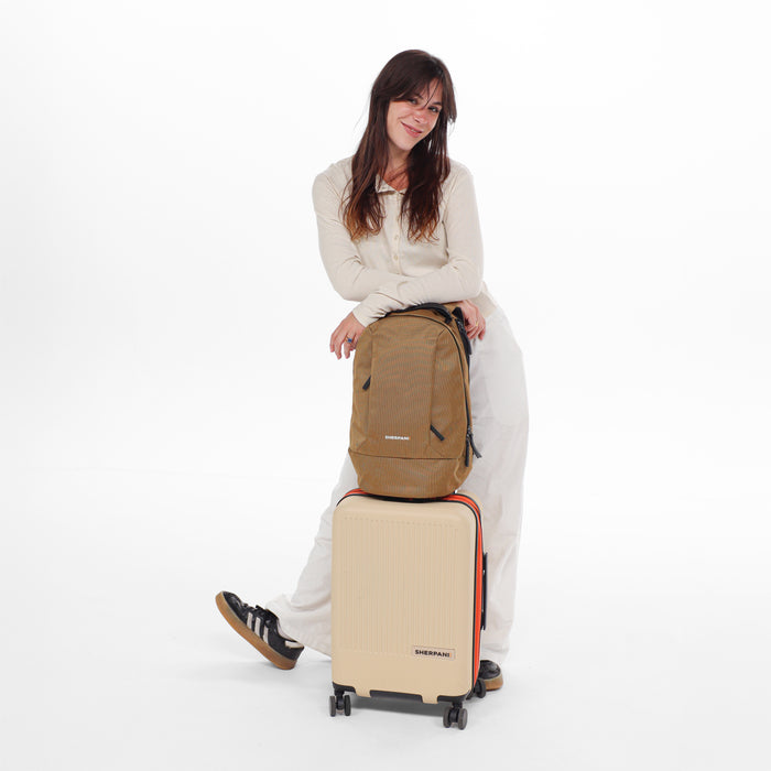 A woman holding a Sherpani Solstice Sand Anti-Theft Travel Backpack on top of a Sherpani Meridian Luggage. #color_sand-tan