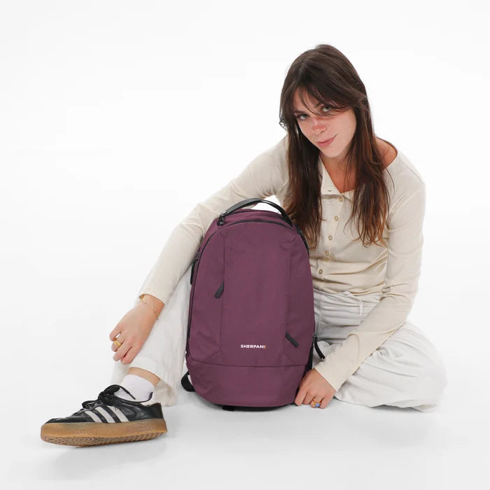 A woman sitting next to a Sherpani Solstice Smoky Orchid Anti-Theft Travel Backpack on her back against a white background.   #color_smoky-orchid-pink