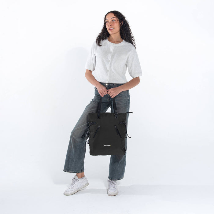 Woman holding a Sherpani Tempest Carbon Black Convertible Tote by tote straps against white background.  #color_carbon-black