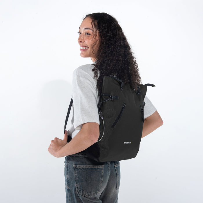 Woman wearing a Sherpani Tempest Carbon Black Convertible Tote on her back against white background. #color_carbon-black