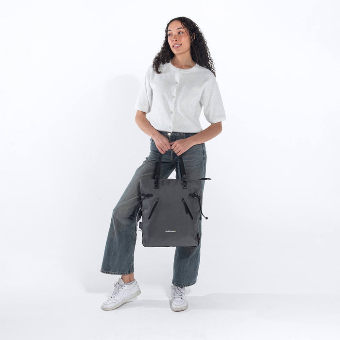 Woman holding a Sherpani Tempest Granite Grey Convertible Tote by tote straps against white background. #color_granite-grey