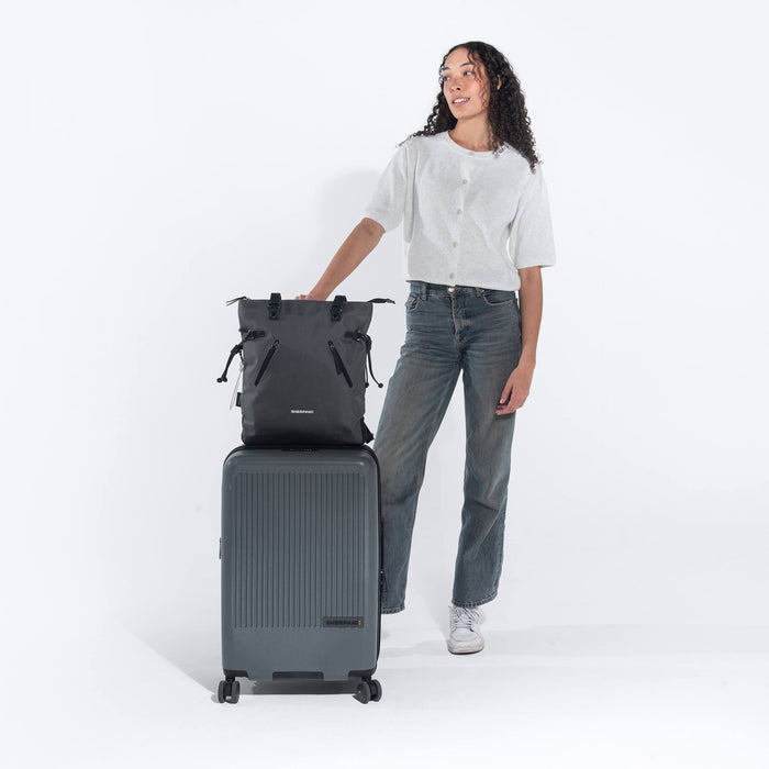 A woman holding a Sherpani Granite Grey Convertible Tote on top Sherpani Meridian Granite Luggage on white backdrop. 
 #color_granite-grey