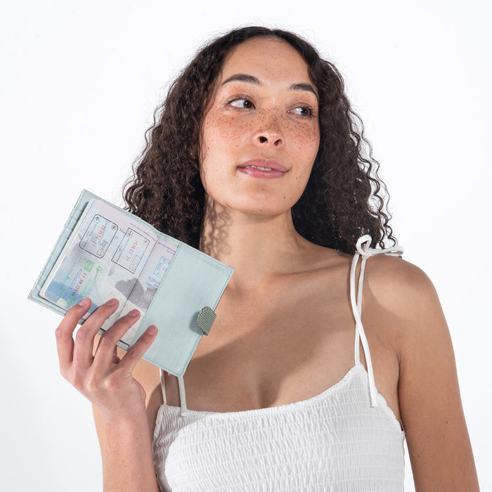 A woman holding a Sherpani Vienna Aloe Mist Green Anti-theft Passport Wallet against a white background.  #color_aloe-mist-green