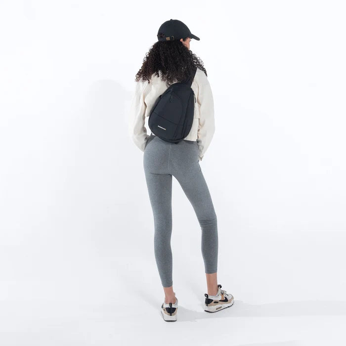 A woman wearing a Sherpani Wayfarer Raven Black Convertible Sling on her back against white background.  #color_raven-black