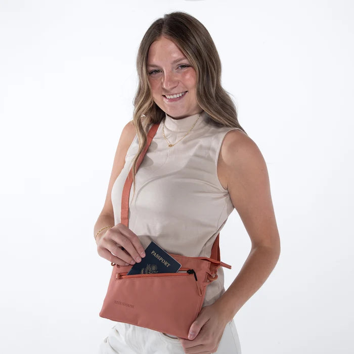 Woman placing a passport into a  Sherpani Zoom Desert Pink Slim RFID Crossbody against white background. #color_desert-pink